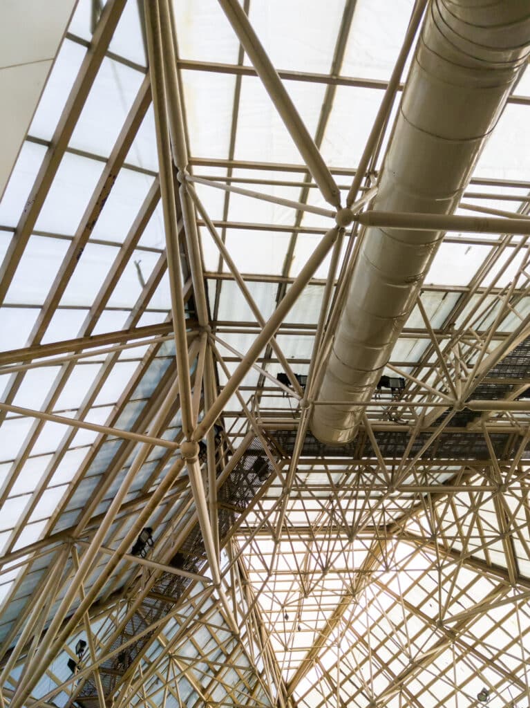 Décapage d'un structure métallique industrielle vue de l'intérieur, avec des poutres et des conduits visibles sous une toiture en verre.