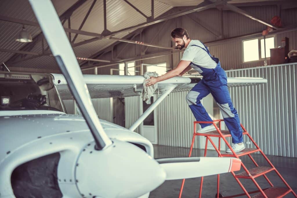 Un technicien en combinaison de travail bleue effectue un décapage par aérogommage sur l'aile d'un avion léger dans un hangar.